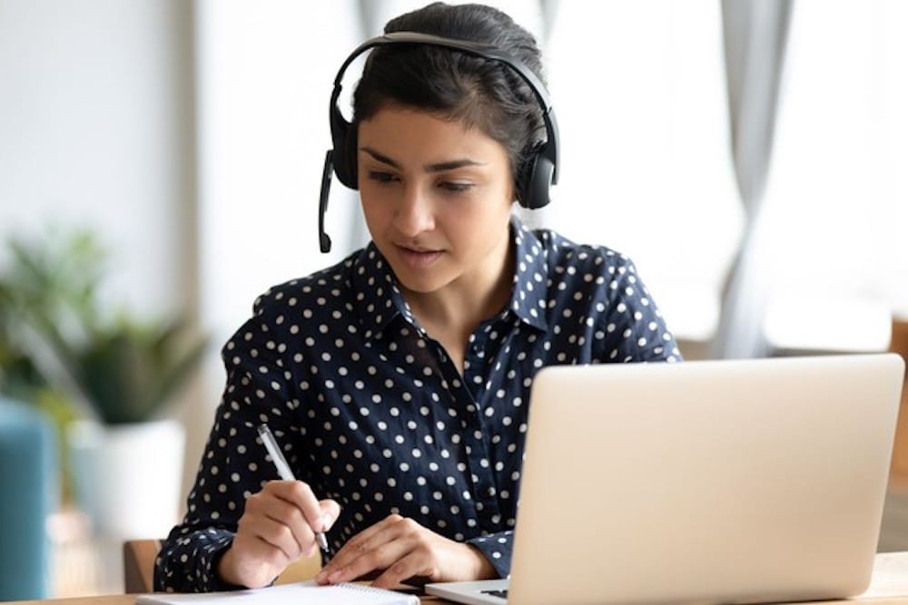 Focused Indian girl wearing headset writing notes, studying online Staff Training