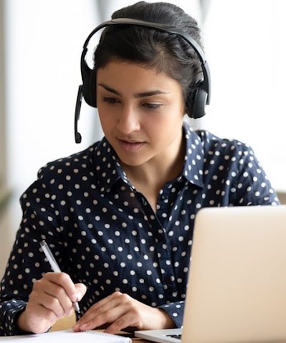 Focused Indian girl wearing headset writing notes, studying online
